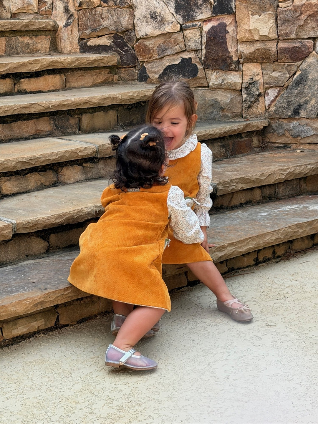 a toddler laughing on stone steps while the little one climbs up beside her, both in rust corduroy jumpers